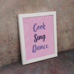 Infidu Cook Sing Dance Kitchen Wall Art with bold navy-blue text on soft pink background, framed in black, modern and lively kitchen design. displayed on the wall in a white frame