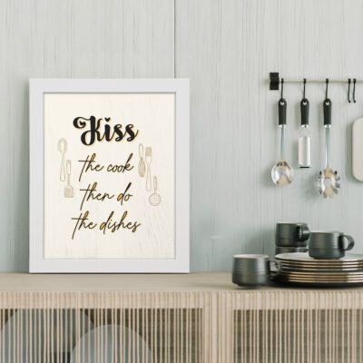 Kiss the Cook Then Do The Dishes wall art with black and gold text, featuring utensil illustrations on a cream background for elegant kitchen decor. displayed on the table