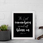 The Lord Remembers Us And Will Bless Us - Psalm 115:12 Bible Verse Wall Art on a black background with white text. displayed on the wall in a white frame