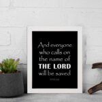 Acts 2:21 Bible wall art featuring bold white text on a black background, reading And everyone who calls on the name of the Lord will be saved. displayed on the wall in a white frame