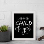 Black wall art with white text reading 'I am a CHILD of god' from John 1:12, showcasing a mix of cursive and bold lettering. displayed on the wall in a white frame