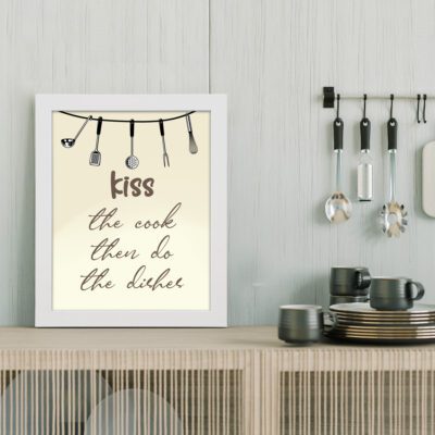 Kiss the Cook Then Do the Dishes kitchen wall art with brown cursive text on beige background and black kitchen utensils illustration. displayed on the table