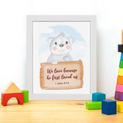 A cute white bear holding a wooden sign with the verse We Love Because He First Loved Us - 1 John 4:19 against a blue sky with clouds. displayed on the table in a white frame