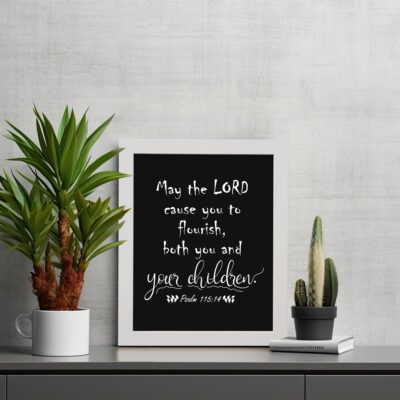 Wall art featuring the quote 'May the Lord cause you to flourish, both you and your children' from Psalm 115:14 on a black background. displayed on the table in a white frame