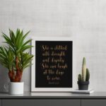 Proverbs 31:25 Christian Wall Art - She Is Clothed With Strength And Dignity, Gold Text on Black Background displayed on the table in a white frame