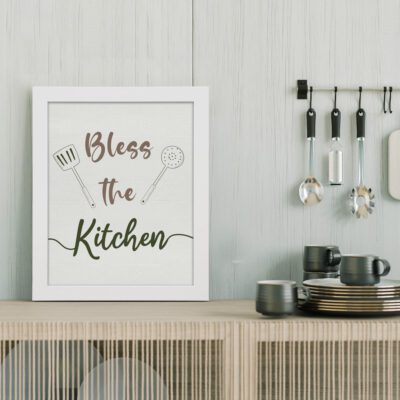 Infidu Bless This Kitchen Kitchen Wall Art with brown and green fonts and kitchen utensil illustrations on a gray background. displayed on the table in a white frame