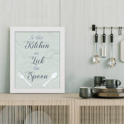 In This Kitchen We Lick The Spoon Kitchen wall art featuring decorative text on a light grey background with fork and spoon illustrations. displayed on the table