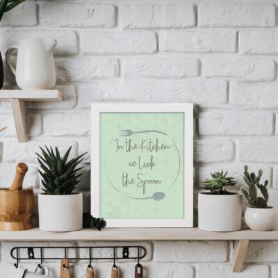 In This Kitchen We Lick The Spoon kitchen wall art with light green background and subtle kitchen utensil pattern in a casual handwritten font. displayed on the table