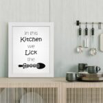 In This Kitchen We Lick the Spoon kitchen wall art featuring black text on a silver-gray background with a minimalist spoon illustration. displayed on the table in a white frame