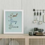 Cook Sing Dance kitchen wall art with hand-drawn fonts, colorful doodles, and a textured light blue background. displayed on the table in a white frame