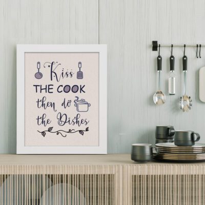 Kitchen wall art with the phrase "Kiss the Cook Then Do the Dishes" in purple and black cursive fonts on a beige background. displayed on the table in white frame