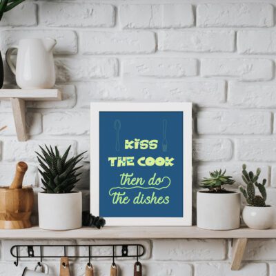 Kiss the cook then do the dishes kitchen wall art with vibrant yellow and green fonts, utensil illustrations, and a deep blue background. displayed on the table in white frame