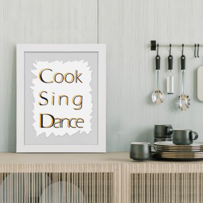 Cook Sing Dance kitchen wall art with black and orange text on a light gray background with jagged white accents. displayed on the table in a white frame