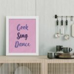 Infidu Cook Sing Dance Kitchen Wall Art with bold navy-blue text on soft pink background, framed in black, modern and lively kitchen design. displayed on the table