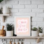 Cook Sing Dance Kitchen Wall Art with pastel pink, green, and blue text on a peach background, perfect for cheerful kitchen decor. displayed on the table in a white frame displayed on the table