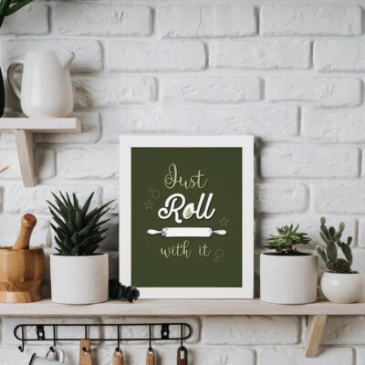 Just Roll With It kitchen wall art with white cursive text, rolling pin illustration, and star patterns on a dark green background. displayed on the table