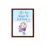 Wall art featuring a joyful bear in winter clothes holding a heart, with the verse We love because He first loved us from 1 John 4:19. hung on the wall in a brown frame