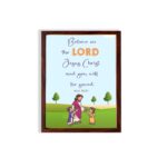 Wall art with a green field, trees, and cartoon figures of Jesus and children, featuring the text 'Believe in the Lord Jesus Christ and you will be saved. hung on the wall in a brown frame