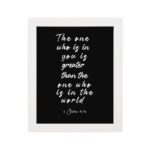 Black wall art with white text displaying The one who is in you is greater than the one who is in the world 1 John 4:4. hung on the wall in a white frame