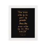 1 John 4:4 Bible verse wall art in gold text on a black background, featuring the quote "The one who is in you is greater than the one who is in the world. hung on the wall in a white frame