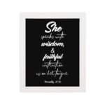 Proverbs 31:26 Bible wall art with white text on a plain black background, featuring cursive and regular fonts emphasizing "She" and "wisdom." hung on the wall in a white frame