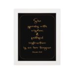 Proverbs 31:26 Bible wall art with gold text on a black background, framed by a golden border. Featuring elegant cursive font. hung on the wall in a white frame