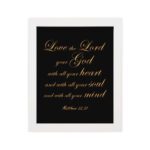 Matthew 22:37 Bible wall art with gold text on a black background, framed in black, reading Love the Lord your God. hung on the wall in a white frame