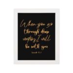 Black wall art with gold cursive text reading When you go through deep waters I will be with you from Isaiah 43:2. hung on the wall in a white frame