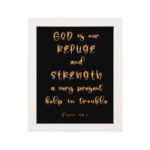 Psalm 46:1 Bible wall art with gold handwritten text on a black background, saying 'God is our refuge and strength. Wall Art hung on the wall in a white frame