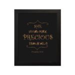 Black wall art with gold text saying SHE IS FAR MORE PRECIOUS THAN JEWELS from Proverbs 31:10, styled with decorative elements. hung on the wall in a black frame