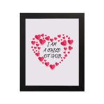 Infidu I Am A Child Of God Bible Wall Art with pink hearts and mixed cursive and print text on a light pink background Wall Art hung on the wall in a black frame