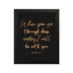 Black wall art with gold cursive text reading When you go through deep waters I will be with you from Isaiah 43:2. hung on the wall in a black frame