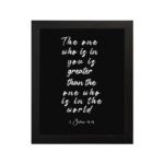 Black wall art with white text displaying The one who is in you is greater than the one who is in the world 1 John 4:4. hung on the wall in a black frame