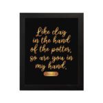 Infidu wall art with the text Like clay in the hand of the potter, so are you in my hand in golden letters on a black background. hung on the wall in a black frame