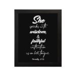 Proverbs 31:26 Bible wall art with white text on a plain black background, featuring cursive and regular fonts emphasizing "She" and "wisdom." hung on the wall in a black frame