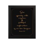 Proverbs 31:26 Bible wall art with gold text on a black background, framed by a golden border. Featuring elegant cursive font. hung on the wall in a black frame