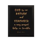 Psalm 46:1 Bible wall art with gold handwritten text on a black background, saying 'God is our refuge and strength. Wall Art hung on the wall in a black frame