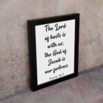 The Lord Of Hosts Is With Us - Psalm 46:7 Bible Verse Wall Art with White Text on Light Background Framed in Black Wall Art displayed on the wall in a black frame