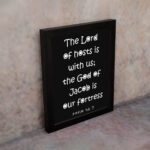 The Lord Of Hosts Is With Us - Psalm 46:7 Bible Verse Wall Art with White Text on Black Background Wall Art displayed on the wall in a black frame