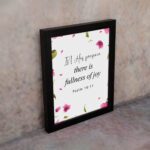 Psalm 16:11 wall art with a white background and pink flower petals, featuring the text 'In His presence there is fullness of joy. Wall Art displayed on the wall in a black frame