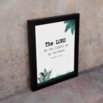 The Lord My God Lights Up My Darkness Psalm 18:28 Bible Verse Wall Art with black text and green tropical leaves on a white background. Wall Art displayed on the wall in a black frame