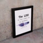 The Lord My God Lights Up My Darkness Psalm 18:28 Bible Verse Wall Art with black text and a purple and blue feather on a white background. Wall Art displayed on the wall in a black frame