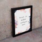 Trust In The Lord And Do Good Psalm 37:3 Bible Verse Wall Art with watercolor and floral designs on a white background. Wall Art displayed on the wall in a black frame