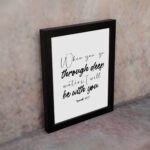 White wall art with black text in script and bold fonts quoting When you go through deep waters, I will be with you from Isaiah 43:2. displayed on the wall in a black frame