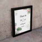 Trust in the Lord and Do Good Psalm 37:3 Bible Wall Art with botanical green leaves and pink flowers on a white background. Wall Art displayed on the wall in a black frame