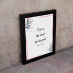 Infidu Bible wall art featuring Psalm 37:3 in black text with blue and pink floral accents and a soft pink border. Wall Art displayed on the wall in a black frame