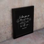 Black wall art with white text and small flower designs quoting When you go through deep waters I will be with you from Isaiah 43:2. displayed on the wall in a black frame