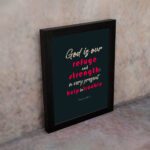 Infidu Psalm 46:1 Bible Wall Art with a deep blue background, pink and white text, featuring God is our refuge and strength. Wall Art displayed on the wall in a black frame