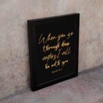 Black wall art with gold cursive text reading When you go through deep waters I will be with you from Isaiah 43:2. displayed on the wall in a black frame