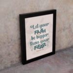 Christian wall art with greenish-blue 'Faith' and 'Fear' text on a light peach floral background. Wall Art displayed on the wall in a black frame